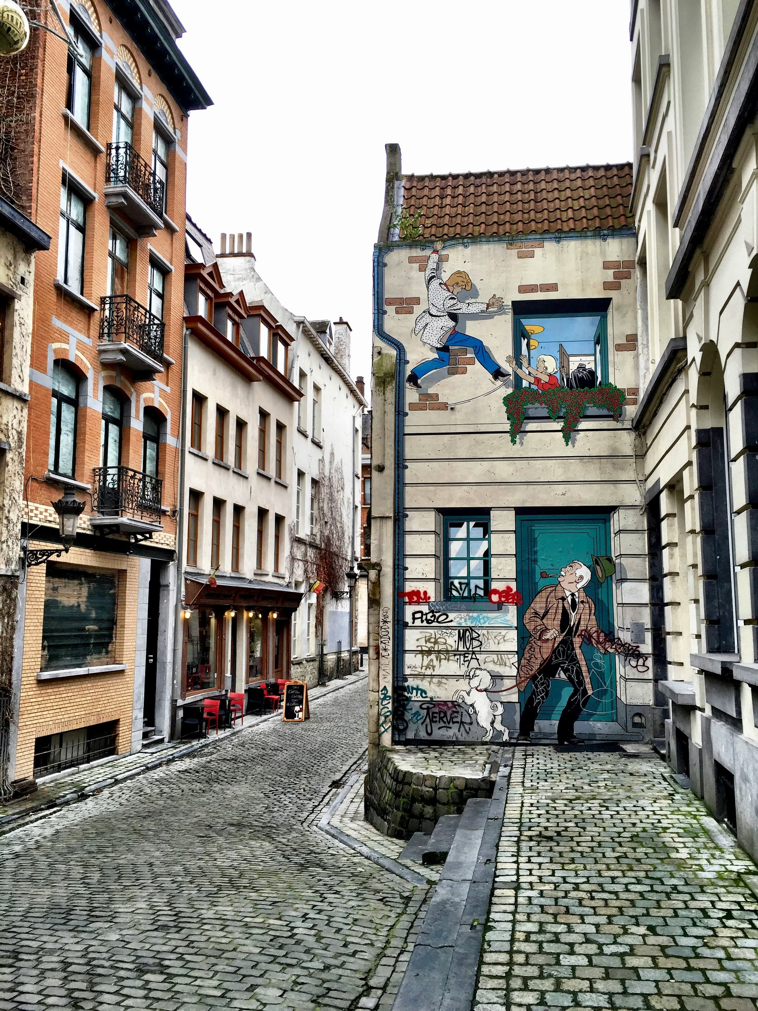 Large comic strip mural on a building side along a narrow, empty cobblestone street.