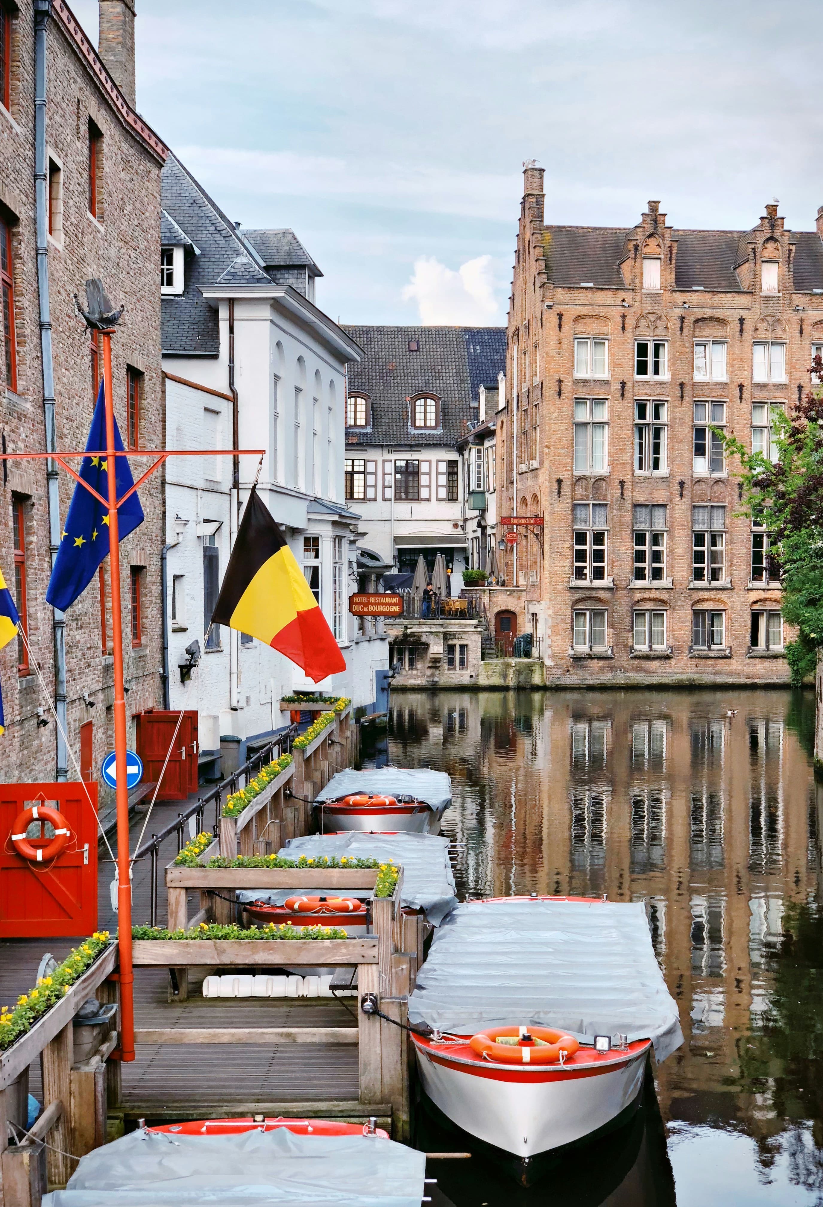 Scenic canal in Bruges, Belgium, featuring docked tour boats, historic brick architecture, and flags.