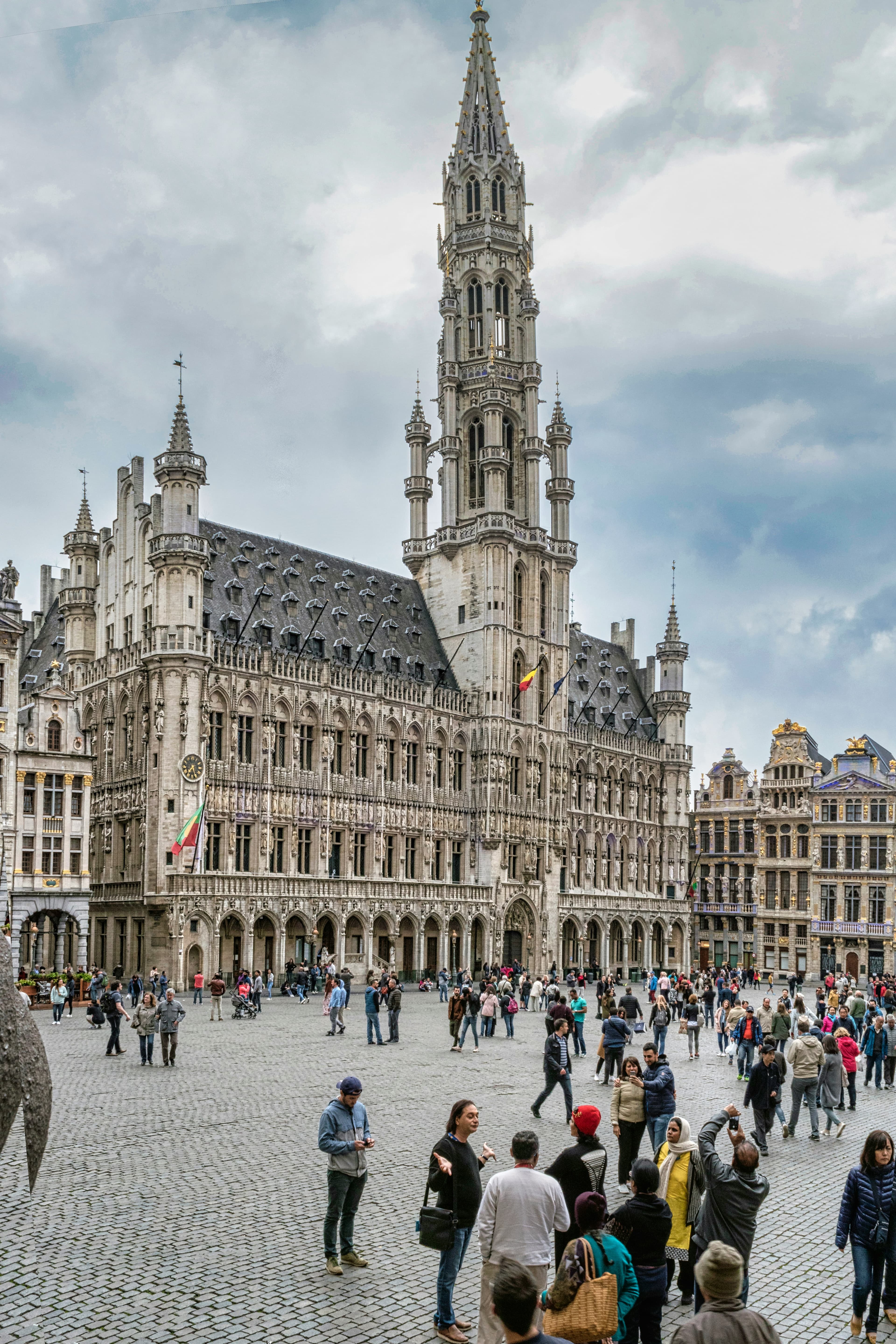 Ornate Gothic Brussels Town Hall overlooks a crowded cobblestone square under a cloudy sky.